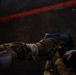 A person in tactical/military gear and camouflage uniform is firing a pistol with a distinctive blue slide in a dark indoor environment. A spent brass casing is visible in the air, indicating the weapon has just been discharged. A second person in tactical gear and helmet is visible in the background.
