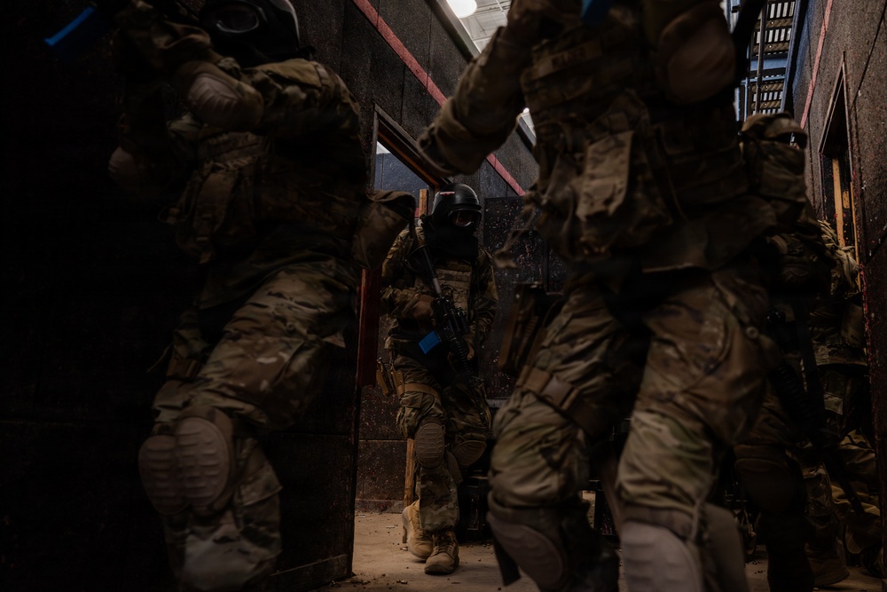 The image shows multiple personnel in full tactical/military gear moving through a narrow corridor or building interior. At least one individual is wearing a ballistic helmet with face protection and carrying a firearm. The scene appears to depict a tactical training exercise or operational building clearance, indicated by blue training markers visible on equipment.