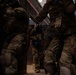 The image shows multiple personnel in full tactical/military gear moving through a narrow corridor or building interior. At least one individual is wearing a ballistic helmet with face protection and carrying a firearm. The scene appears to depict a tactical training exercise or operational building clearance, indicated by blue training markers visible on equipment.