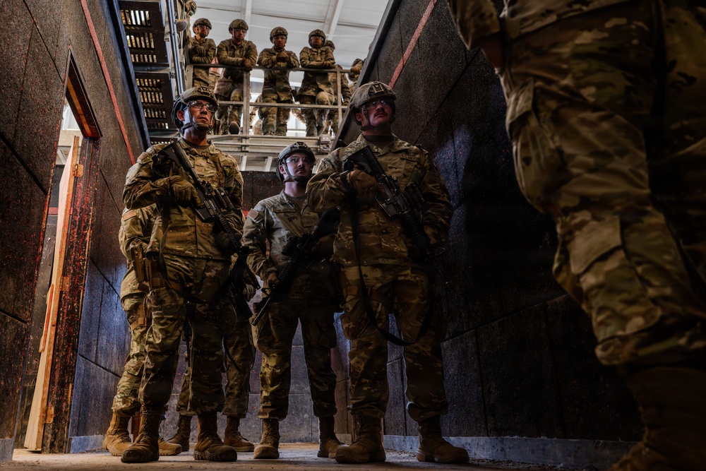 A group of military personnel in full combat gear are gathered in what appears to be a training facility or urban operations building. Several soldiers stand in a lower level while additional personnel observe from an elevated platform or mezzanine above. The scene strongly suggests a military training exercise, likely focused on close-quarters battle or urban warfare tactics.