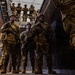 A group of military personnel in full combat gear are gathered in what appears to be a training facility or urban operations building. Several soldiers stand in a lower level while additional personnel observe from an elevated platform or mezzanine above. The scene strongly suggests a military training exercise, likely focused on close-quarters battle or urban warfare tactics.
