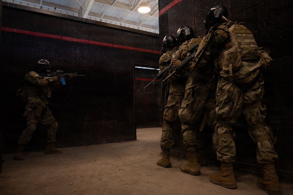 The image depicts a military or law enforcement training exercise taking place inside an indoor close-quarters battle (CQB) training facility. Four personnel in full tactical gear, camouflage uniforms, and protective helmets are conducting a room-clearing or breach drill. One operator is in a shooting stance with a rifle raised, while three others are stacked against a dark wall awaiting entry.