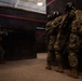 The image depicts a military or law enforcement training exercise taking place inside an indoor close-quarters battle (CQB) training facility. Four personnel in full tactical gear, camouflage uniforms, and protective helmets are conducting a room-clearing or breach drill. One operator is in a shooting stance with a rifle raised, while three others are stacked against a dark wall awaiting entry.