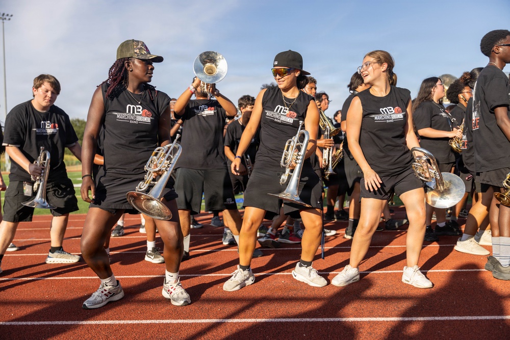 A high school or college marching band rehearses on a red running track during what appears to be an outdoor practice session. Members are wearing matching black sleeveless shirts reading 'Marching Bobcat Band' (MB) and carrying brass instruments including trumpets and flugelhorns. The atmosphere appears energetic and social, with several members smiling and interacting.