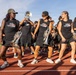 A high school or college marching band rehearses on a red running track during what appears to be an outdoor practice session. Members are wearing matching black sleeveless shirts reading 'Marching Bobcat Band' (MB) and carrying brass instruments including trumpets and flugelhorns. The atmosphere appears energetic and social, with several members smiling and interacting.