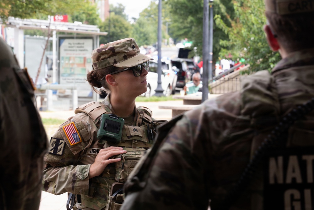 A female soldier in U.S. Army camouflage uniform is engaged in conversation with other personnel in an outdoor urban setting. She is wearing full tactical gear including a plate carrier, sunglasses, and a patrol cap. The text on the right figure's uniform partially reads 'National Guard,' indicating this is a National Guard deployment or operation.