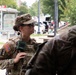 A female soldier in U.S. Army camouflage uniform is engaged in conversation with other personnel in an outdoor urban setting. She is wearing full tactical gear including a plate carrier, sunglasses, and a patrol cap. The text on the right figure's uniform partially reads 'National Guard,' indicating this is a National Guard deployment or operation.