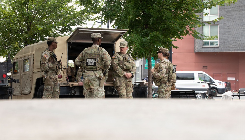 Four National Guard soldiers in OCP (Operational Camouflage Pattern) uniforms are gathered near the open rear of a tan military HMMWV (Humvee) in an urban setting. They appear to be in conversation or conducting a briefing near the vehicle. The scene takes place in what appears to be a city plaza or parking area with residential/commercial buildings visible in the background.