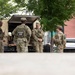Four National Guard soldiers in OCP (Operational Camouflage Pattern) uniforms are gathered near the open rear of a tan military HMMWV (Humvee) in an urban setting. They appear to be in conversation or conducting a briefing near the vehicle. The scene takes place in what appears to be a city plaza or parking area with residential/commercial buildings visible in the background.