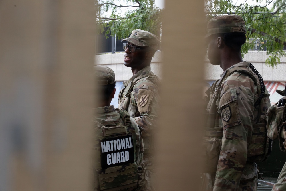 The image shows multiple National Guard soldiers in military camouflage uniforms photographed through what appears to be a fence or barrier. At least three soldiers are visible, with one clearly displaying a 'NATIONAL GUARD' patch on their tactical vest. The soldiers appear to be standing and conversing in an outdoor urban setting.