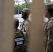 The image shows multiple National Guard soldiers in military camouflage uniforms photographed through what appears to be a fence or barrier. At least three soldiers are visible, with one clearly displaying a 'NATIONAL GUARD' patch on their tactical vest. The soldiers appear to be standing and conversing in an outdoor urban setting.
