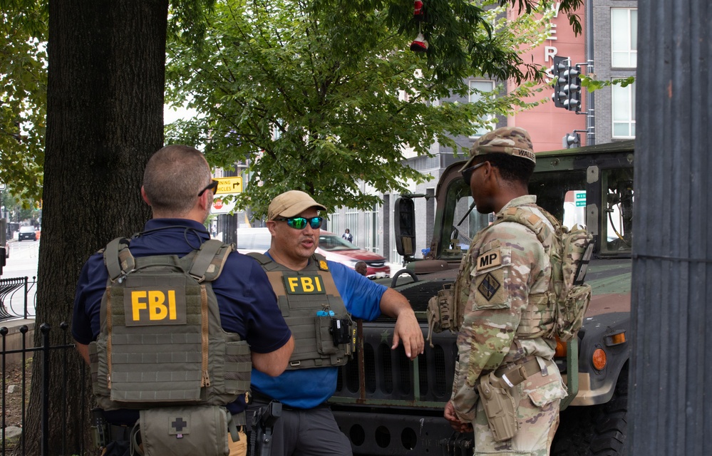 Two FBI agents wearing tactical vests labeled 'FBI' are conversing with a U.S. Army Military Police (MP) soldier in full camouflage uniform on an urban street. A military Humvee is visible in the background, suggesting a joint law enforcement and military operation or security detail. The setting appears to be a city street with trees, traffic signals, and commercial buildings visible.