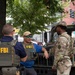 Two FBI agents wearing tactical vests labeled 'FBI' are conversing with a U.S. Army Military Police (MP) soldier in full camouflage uniform on an urban street. A military Humvee is visible in the background, suggesting a joint law enforcement and military operation or security detail. The setting appears to be a city street with trees, traffic signals, and commercial buildings visible.