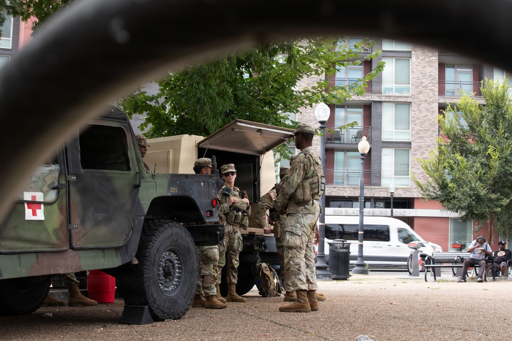Military personnel in camouflage uniforms are gathered around a military HMMWV (Humvee) marked with a Red Cross symbol, indicating a medical vehicle, in what appears to be an urban public plaza or park. The photo is taken from a low angle through what appears to be a bicycle wheel frame, giving a partially obstructed perspective. Civilians can be seen seated on benches in the background near a modern multi-story building.