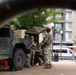 Military personnel in camouflage uniforms are gathered around a military HMMWV (Humvee) marked with a Red Cross symbol, indicating a medical vehicle, in what appears to be an urban public plaza or park. The photo is taken from a low angle through what appears to be a bicycle wheel frame, giving a partially obstructed perspective. Civilians can be seen seated on benches in the background near a modern multi-story building.