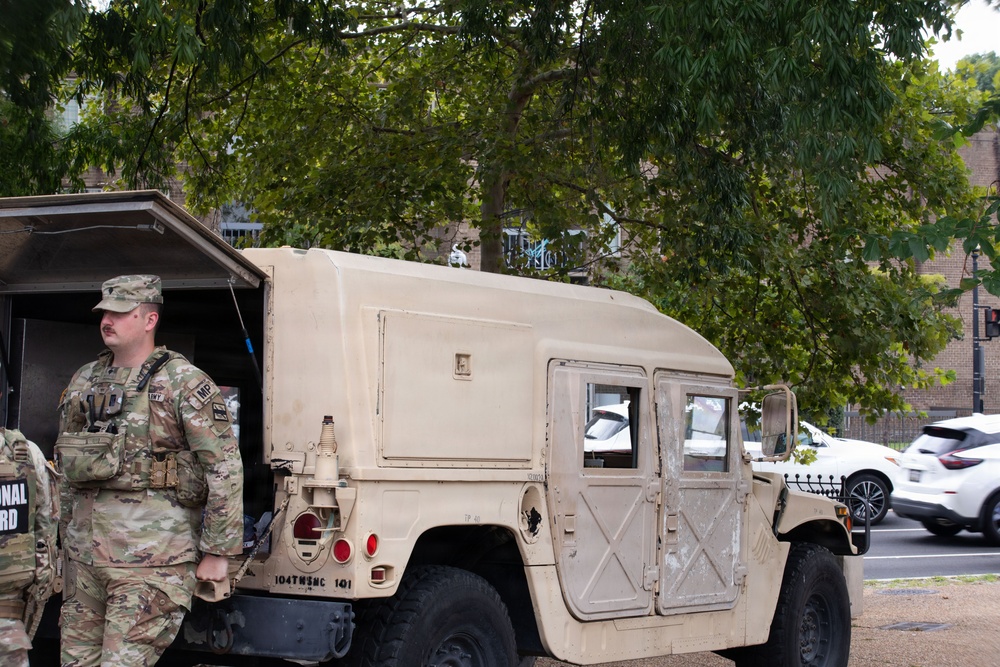 The image shows U.S. Army National Guard soldiers standing near a tan/desert-colored HMMWV (Humvee) military vehicle on an urban street. At least one soldier is clearly visible wearing OCP (Operational Camouflage Pattern) uniform with an 'MP' (Military Police) patch and tactical vest. Partial text on a banner in the background reads 'NATIONAL GUARD'.