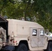 The image shows U.S. Army National Guard soldiers standing near a tan/desert-colored HMMWV (Humvee) military vehicle on an urban street. At least one soldier is clearly visible wearing OCP (Operational Camouflage Pattern) uniform with an 'MP' (Military Police) patch and tactical vest. Partial text on a banner in the background reads 'NATIONAL GUARD'.