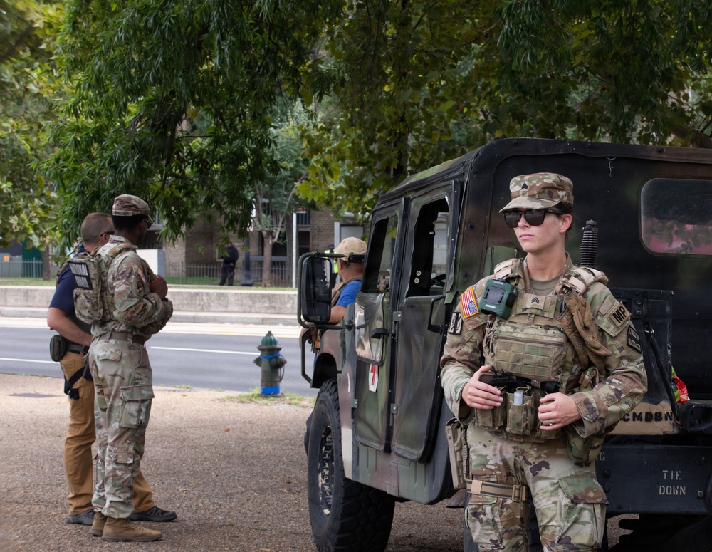 The image shows U.S. military personnel, including at least one Military Police (MP) soldier, stationed near a military Humvee on what appears to be a public street or park area. The soldiers are in full combat/duty gear including multicam uniforms, tactical vests, and equipment. The setting appears to be an urban environment, possibly during a domestic deployment or security operation.