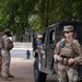 The image shows U.S. military personnel, including at least one Military Police (MP) soldier, stationed near a military Humvee on what appears to be a public street or park area. The soldiers are in full combat/duty gear including multicam uniforms, tactical vests, and equipment. The setting appears to be an urban environment, possibly during a domestic deployment or security operation.