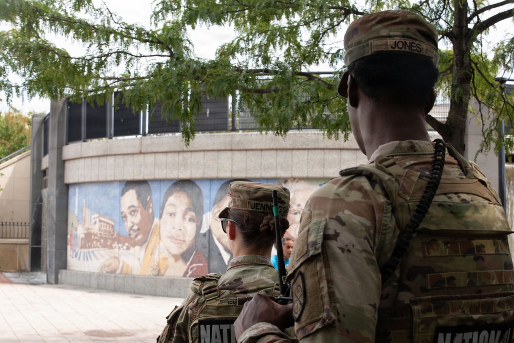 Two National Guard soldiers in camouflage uniforms are standing near a public mural depicting children's faces. The soldiers, identified by name tapes as 'Jones' and 'Jennings', are facing away from the camera toward the mural and surrounding area. The scene appears to depict a security or civil support mission in an urban public space.