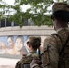 Two National Guard soldiers in camouflage uniforms are standing near a public mural depicting children's faces. The soldiers, identified by name tapes as 'Jones' and 'Jennings', are facing away from the camera toward the mural and surrounding area. The scene appears to depict a security or civil support mission in an urban public space.