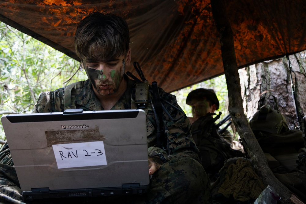 A military personnel wearing camouflage face paint and MARPAT uniform operates a ruggedized Panasonic laptop labeled 'RAV 2-3' under a camouflage tarp shelter in a wooded environment. A second similarly equipped service member is visible in the background. The setting appears to be a field training exercise or tactical field operation.