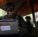A military personnel wearing camouflage face paint and MARPAT uniform operates a ruggedized Panasonic laptop labeled 'RAV 2-3' under a camouflage tarp shelter in a wooded environment. A second similarly equipped service member is visible in the background. The setting appears to be a field training exercise or tactical field operation.