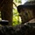 Multi-Domain Reconnaissance Course| Reconnaissance and Surveillance Exercise A military personnel member is positioned in a wooded environment wearing full camouflage gear including face paint and a boonie hat. The individual appears to be in a surveillance or observation posture, holding a weapon with an optical scope attachment. The image is taken from a low angle in dappled forest lighting, emphasizing concealment and tactical positioning.