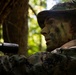 A military personnel member is positioned in a wooded environment wearing full camouflage gear including face paint and a boonie hat. The individual appears to be in a surveillance or observation posture, holding a weapon with an optical scope attachment. The image is taken from a low angle in dappled forest lighting, emphasizing concealment and tactical positioning.