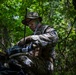 A military personnel in full camouflage uniform and face paint is operating what appears to be a tactical radio or communications device in a wooded, forested environment. The individual is wearing MARPAT (Marine pattern) digital camouflage, suggesting affiliation with the U.S. Marine Corps. The setting and equipment suggest a field training exercise or operational deployment.