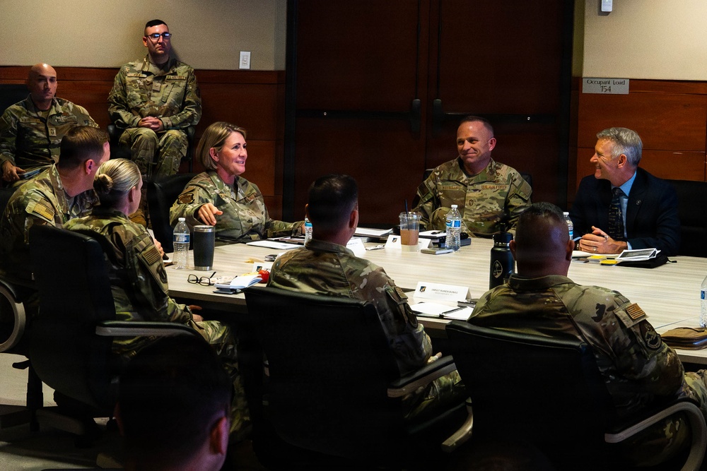 A military briefing or roundtable meeting is taking place in a conference room, with multiple uniformed personnel in OCP (Operational Camouflage Pattern) uniforms seated around a rectangular table. One civilian male in a dark suit and one female officer appear to be central participants in the discussion. The setting appears to be a formal military installation conference room with an occupancy load sign visible on the wall.
