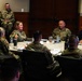 A military briefing or roundtable meeting is taking place in a conference room, with multiple uniformed personnel in OCP (Operational Camouflage Pattern) uniforms seated around a rectangular table. One civilian male in a dark suit and one female officer appear to be central participants in the discussion. The setting appears to be a formal military installation conference room with an occupancy load sign visible on the wall.