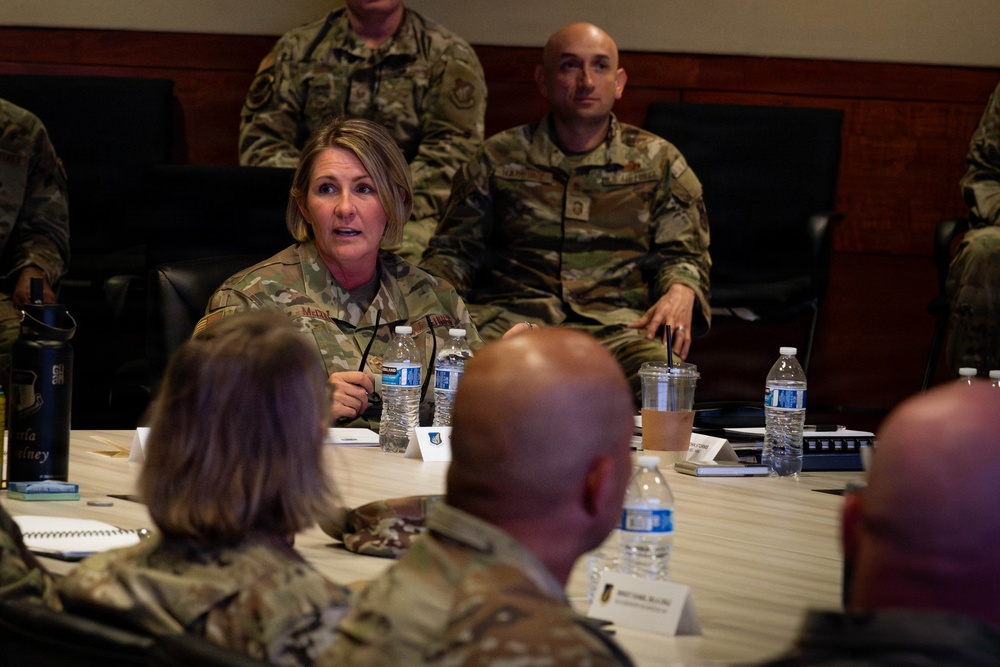 A military briefing or conference is taking place in a formal meeting room, with personnel in U.S. Air Force/Space Force OCP (Operational Camouflage Pattern) uniforms seated around a conference table. A female officer appears to be speaking or presenting to the group. Name placards, water bottles, and documents are visible on the table.