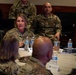 A military briefing or conference is taking place in a formal meeting room, with personnel in U.S. Air Force/Space Force OCP (Operational Camouflage Pattern) uniforms seated around a conference table. A female officer appears to be speaking or presenting to the group. Name placards, water bottles, and documents are visible on the table.