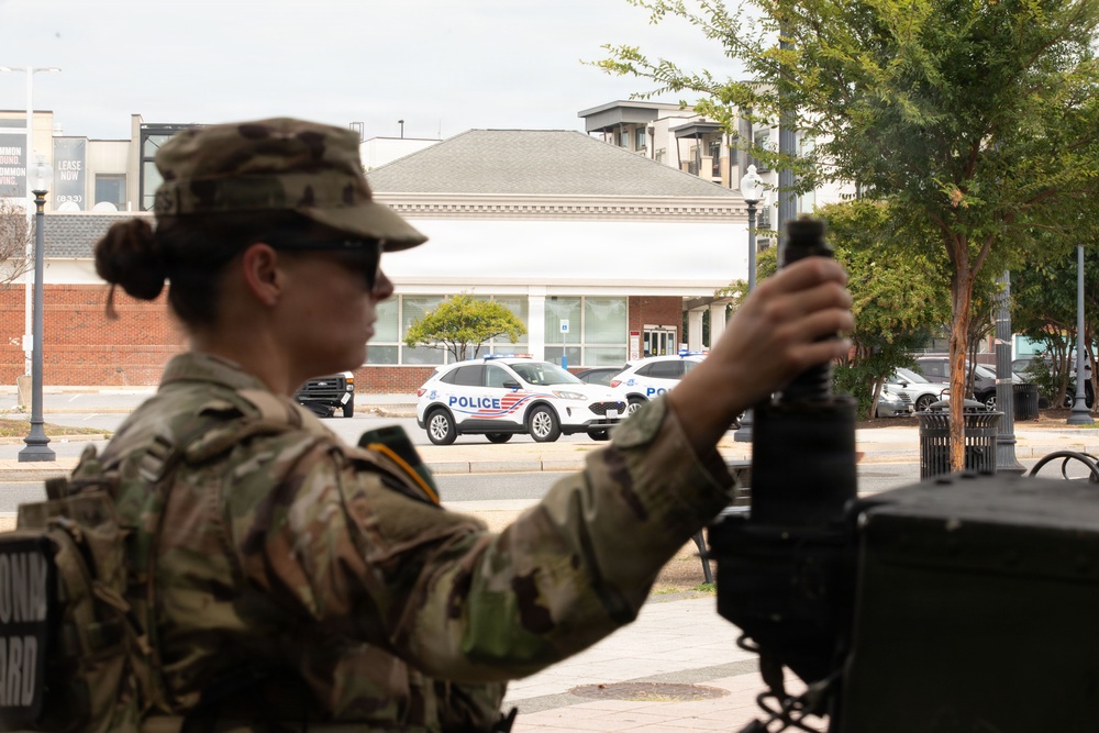 A female soldier in U.S. Army camouflage uniform operates or adjusts equipment mounted on a military vehicle in an urban setting. A Metropolitan Police Department (Washington D.C.) Ford Escape SUV is visible in the background on a city street. Partial text on the soldier's vest reads 'NATIONAL GUARD', indicating this is a National Guard deployment.