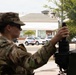 A female soldier in U.S. Army camouflage uniform operates or adjusts equipment mounted on a military vehicle in an urban setting. A Metropolitan Police Department (Washington D.C.) Ford Escape SUV is visible in the background on a city street. Partial text on the soldier's vest reads 'NATIONAL GUARD', indicating this is a National Guard deployment.