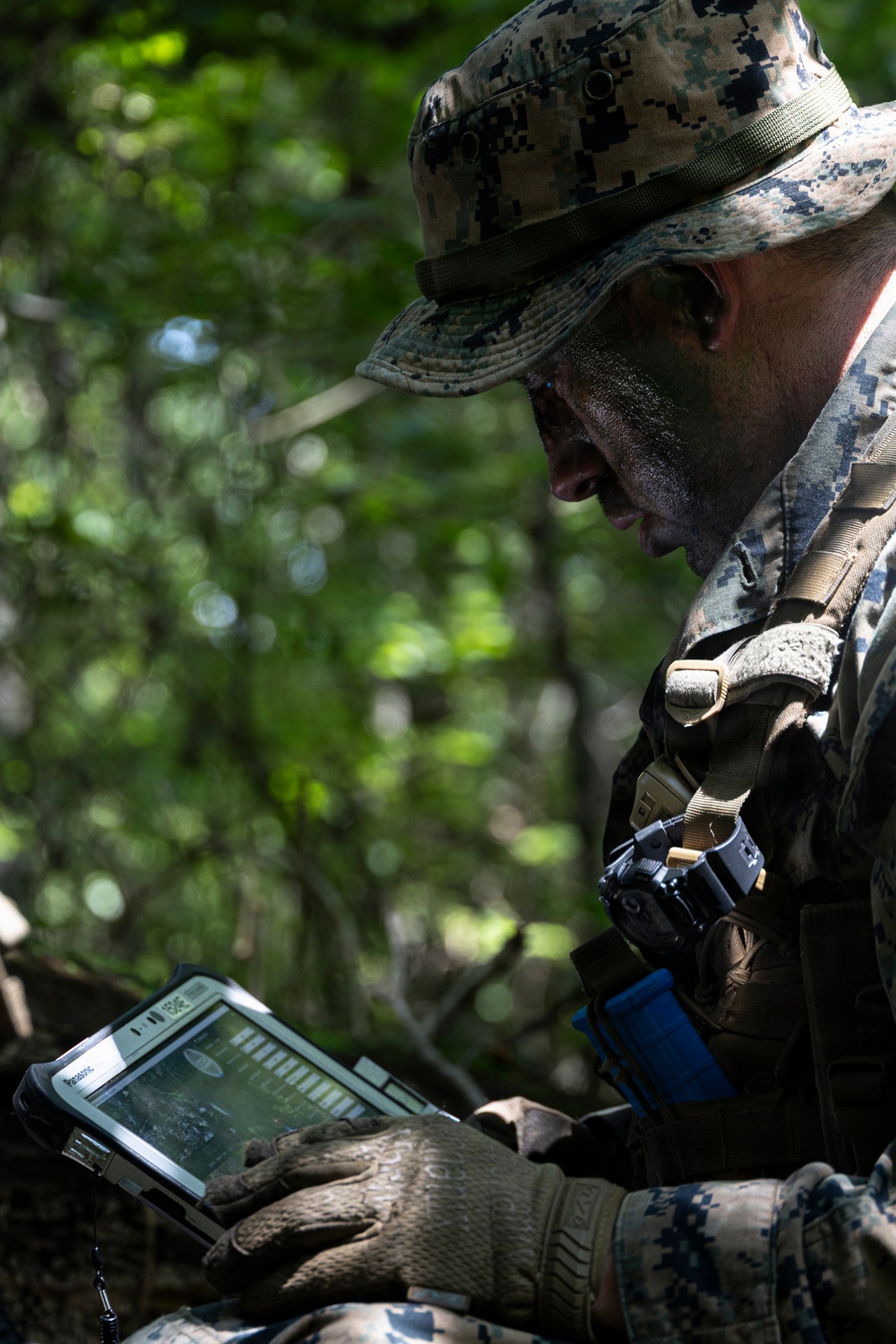 A military personnel wearing MARPAT digital camouflage uniform and boonie hat is operating a ruggedized Panasonic tablet device in a wooded outdoor environment. The individual has camouflage face paint applied and is wearing tactical gloves while reviewing what appears to be a digital map or tactical display. The setting suggests a field training exercise or operational deployment.