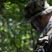 A military personnel wearing MARPAT digital camouflage uniform and boonie hat is operating a ruggedized Panasonic tablet device in a wooded outdoor environment. The individual has camouflage face paint applied and is wearing tactical gloves while reviewing what appears to be a digital map or tactical display. The setting suggests a field training exercise or operational deployment.