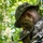 Multi-Domain Reconnaissance Course| Reconnaissance and Surveillance Exercise Two military personnel in woodland camouflage uniforms and face paint are engaged in a field exercise in a forested environment. The foreground individual is wearing a boonie hat and tactical gloves, holding a writing instrument and appearing to record or review information on a small device or notepad. The setting and equipment suggest a land navigation or reconnaissance training exercise.