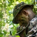Two military personnel in woodland camouflage uniforms and face paint are engaged in a field exercise in a forested environment. The foreground individual is wearing a boonie hat and tactical gloves, holding a writing instrument and appearing to record or review information on a small device or notepad. The setting and equipment suggest a land navigation or reconnaissance training exercise.