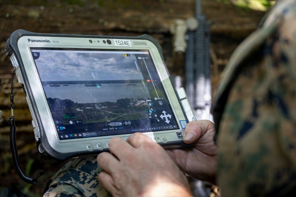 A military personnel in camouflage uniform is operating a Panasonic FZ-G1 Toughpad rugged tablet (unit ID: 1524E) displaying what appears to be aerial drone surveillance footage of a coastal or riverine area. The screen shows a tactical software interface with overlaid navigation markers, targeting indicators, and geographic data. A rifle is visible in the background, suggesting a field operational environment.