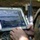 Multi-Domain Reconnaissance Course| Reconnaissance and Surveillance Exercise A military personnel in camouflage uniform is operating a Panasonic FZ-G1 Toughpad rugged tablet (unit ID: 1524E) displaying what appears to be aerial drone surveillance footage of a coastal or riverine area. The screen shows a tactical software interface with overlaid navigation markers, targeting indicators, and geographic data. A rifle is visible in the background, suggesting a field operational environment.