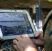 A military personnel in camouflage uniform is operating a Panasonic FZ-G1 Toughpad rugged tablet (unit ID: 1524E) displaying what appears to be aerial drone surveillance footage of a coastal or riverine area. The screen shows a tactical software interface with overlaid navigation markers, targeting indicators, and geographic data. A rifle is visible in the background, suggesting a field operational environment.