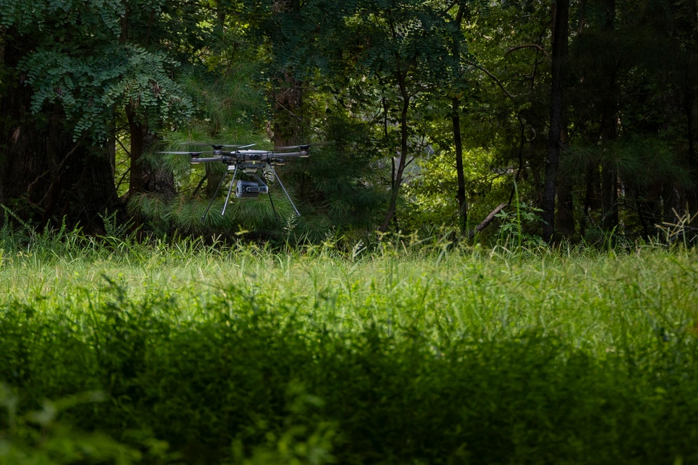 A multi-rotor drone (likely a hexacopter or octocopter) hovers at low altitude above a grassy clearing at the edge of a dense, green forest. The drone is equipped with a payload or camera gimbal suspended beneath its frame. The image is captured from a ground-level perspective, emphasizing the lush foreground vegetation and wooded backdrop.