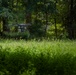A multi-rotor drone (likely a hexacopter or octocopter) hovers at low altitude above a grassy clearing at the edge of a dense, green forest. The drone is equipped with a payload or camera gimbal suspended beneath its frame. The image is captured from a ground-level perspective, emphasizing the lush foreground vegetation and wooded backdrop.