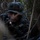 Multi-Domain Reconnaissance Course| Reconnaissance and Surveillance Exercise A military service member in digital camouflage uniform and boonie hat is positioned low in dense jungle or forest vegetation, appearing to be in a tactical concealment position. The individual is holding a weapon and appears alert, scanning their surroundings. The setting suggests a field training exercise or combat patrol in a jungle environment.