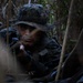 A military service member in digital camouflage uniform and boonie hat is positioned low in dense jungle or forest vegetation, appearing to be in a tactical concealment position. The individual is holding a weapon and appears alert, scanning their surroundings. The setting suggests a field training exercise or combat patrol in a jungle environment.