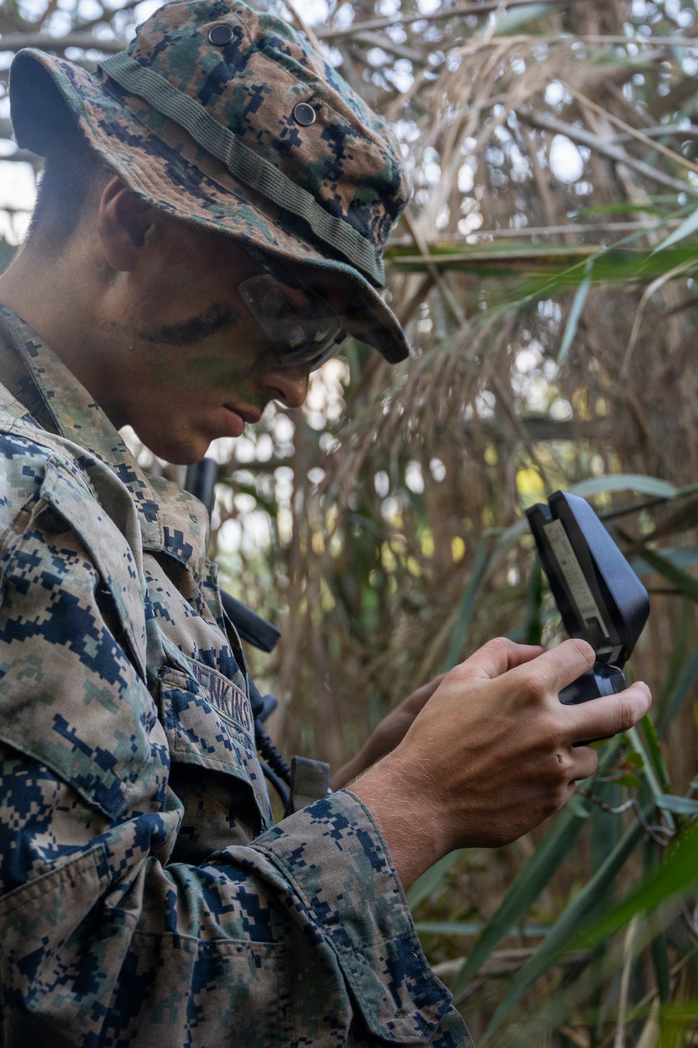 A U.S. Marine in MARPAT digital camouflage uniform and boonie cover is examining a lensatic compass while positioned in dense vegetation. The service member has camouflage face paint applied and a name tape partially visible reading 'ENKINS' (likely Jenkins). The image appears to document a land navigation training exercise in a jungle or heavily vegetated environment.