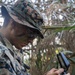 A U.S. Marine in MARPAT digital camouflage uniform and boonie cover is examining a lensatic compass while positioned in dense vegetation. The service member has camouflage face paint applied and a name tape partially visible reading 'ENKINS' (likely Jenkins). The image appears to document a land navigation training exercise in a jungle or heavily vegetated environment.