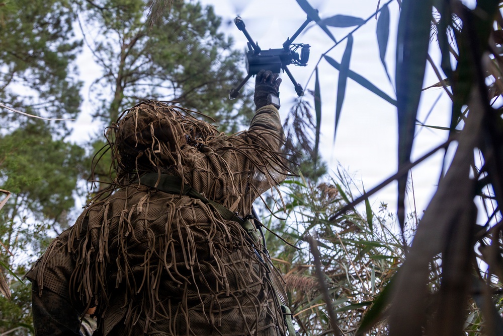 A person wearing a ghillie suit is holding up a small quadcopter drone in an outdoor wooded environment. The individual is photographed from a low angle looking upward, with pine trees and overcast sky visible in the background. The scene suggests a military or tactical reconnaissance context involving unmanned aerial vehicle (UAV) deployment.